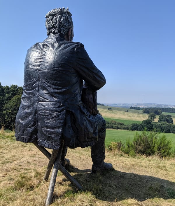 Sculpture of a man sitting down on a stroll, placed on a green hill looking out across the countryside under a blue sky